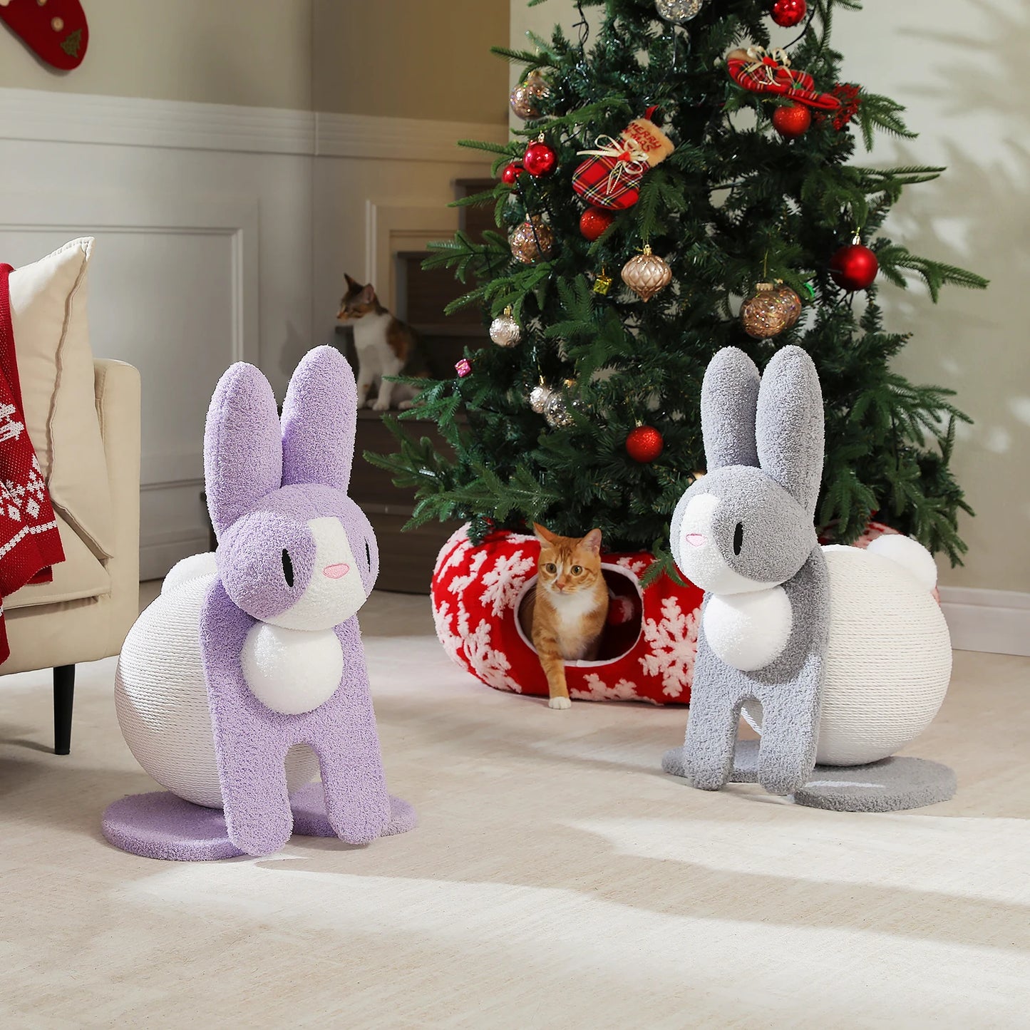 Two bunny-shaped cat scratching posts in front of a decorated Christmas tree with a cat in a red and white striped tunnel.