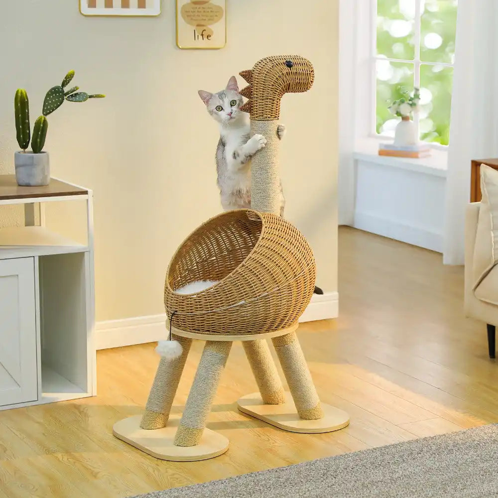Cat playing on a dinosaur-shaped wicker cat tree in a room with a window and plants.